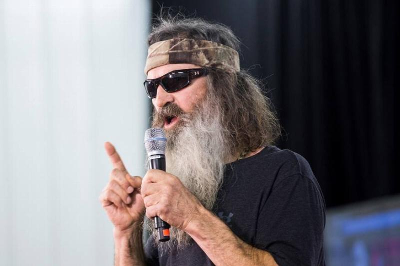 Phil Robertson appears on stage holding a microphone and pointing with his right index finger