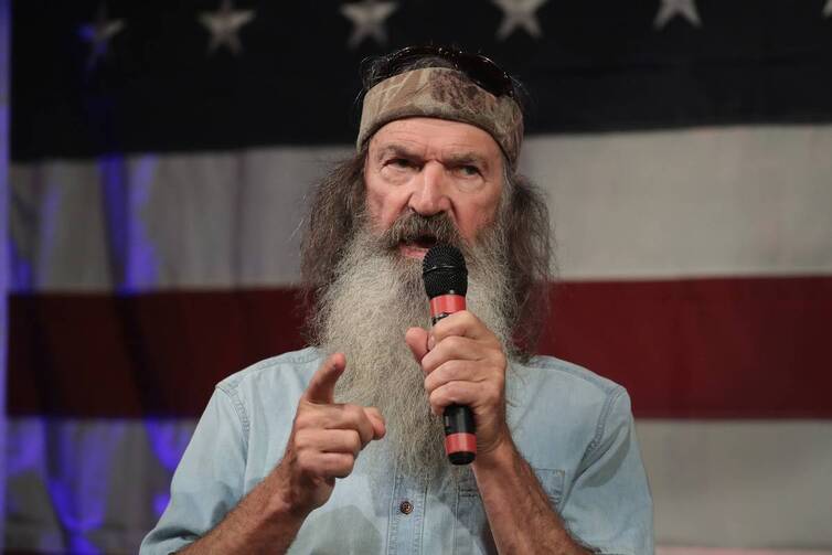Phil Robertson speaking into a microphone in front of an American flag backdrop