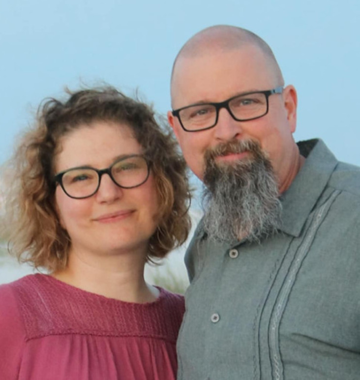 A close-up of a smiling woman with curly hair and glasses, standing next to a bearded man wearing glasses.