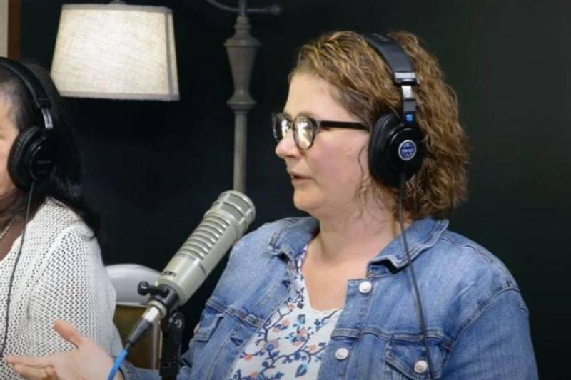 A woman with curly hair and glasses is wearing headphones and speaking into a studio microphone, indicative of a podcast recording session