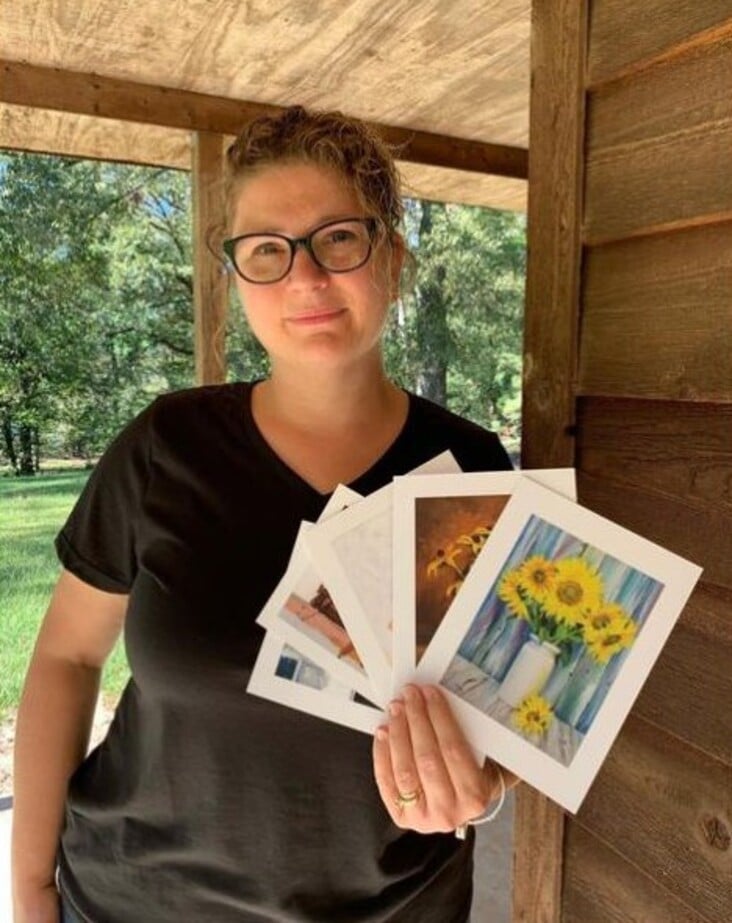 A woman with glasses and curly hair stands under a wooden shelter, holding up several photographs with visible images of sunflowers