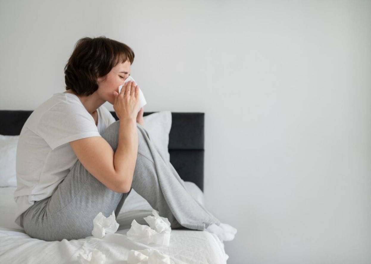 Sick woman sitting in bed blowing her nose surrounded by tissues
