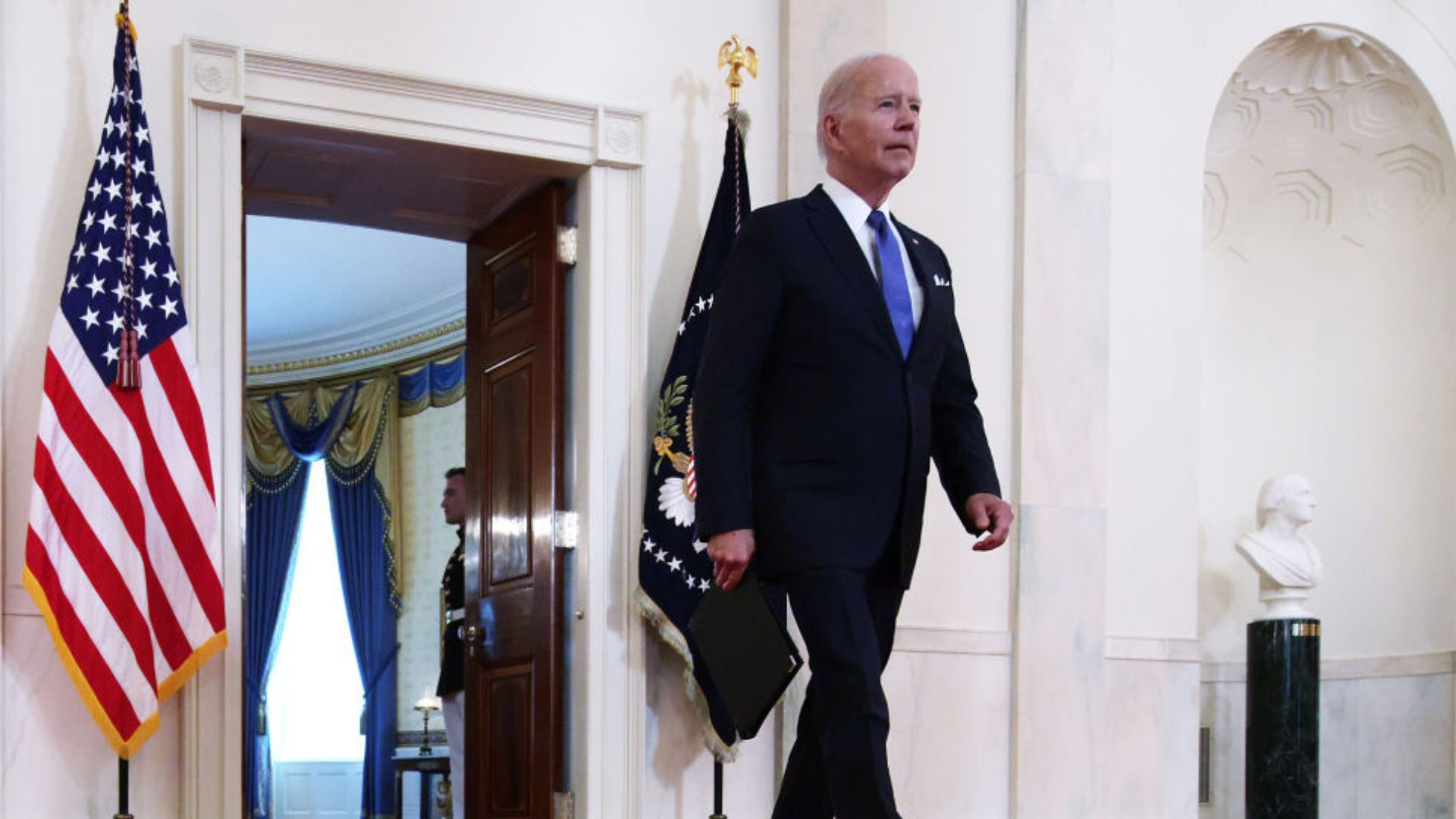 President Joe Biden walking in a grand hallway in The White House, with an American flag to his left, the Presidential seal on a flag next to him
