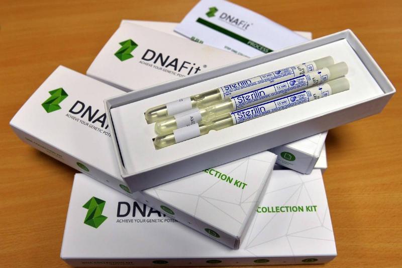 A collection of DNAFit genetic testing kits arranged on a wooden table. One box is open, revealing several test tubes with blue caps inside, while others are closed with the brand's logo visible on the lids