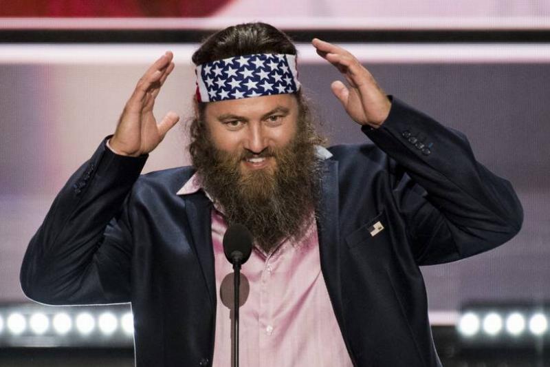 A man with a full beard and long hair wearing an American flag bandana adjusts his headband while speaking into a microphone