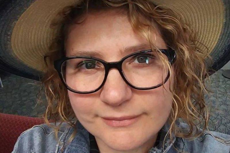 Close-up selfie of a woman with curly hair wearing a straw hat and black-framed glasses