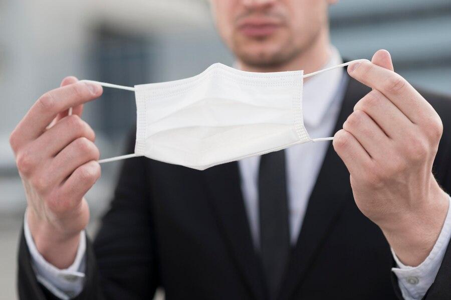 Man in a suit holds a white surgical mask in front of him