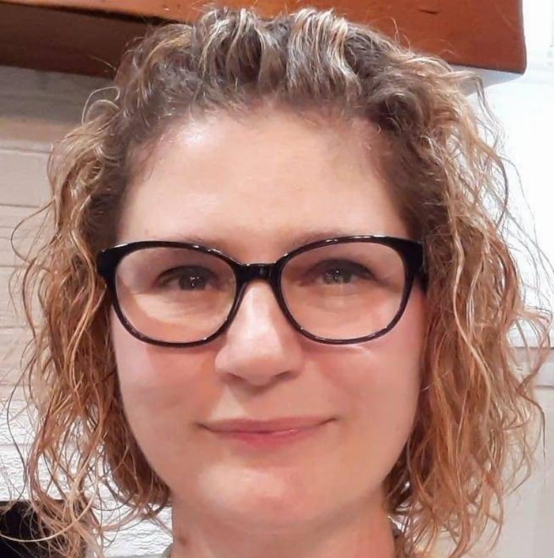 Close-up of a woman with curly hair and black-framed glasses. She has a gentle smile and is looking directly at the camera