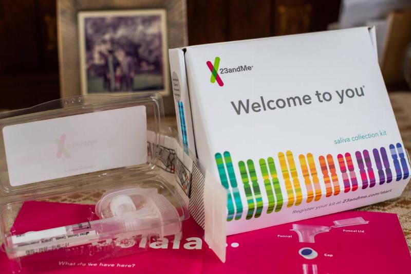 An open 23andMe genetic testing kit is displayed on a table. The kit includes a saliva collection tube, instructions, and a box with the text 'Welcome to you'.