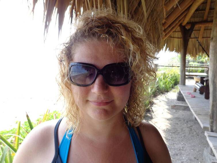 A close up portrait of a woman with blond curly hair wearing sunglasses in a tropical setting