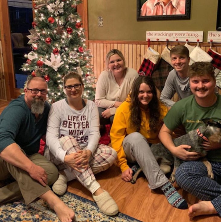 A family of five, with three young adults, sits smiling in front of a decorated Christmas tree